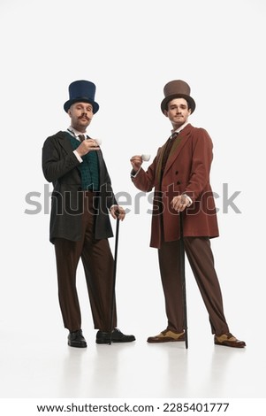 Morning drink. Two gentlemen wearing old vintage suit and hat standing with cup of coffee and looking at camera over white background. Concept of historical remake, comparison of eras, retro, vintage