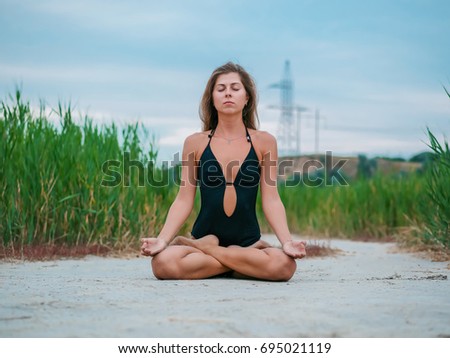 Morning exercise. Lotus pose. Woman stretching hip, hamstring muscles and groin area. Fit fitness athlete girl exercising sports stretches in black swimsuit at sunset near reeds.