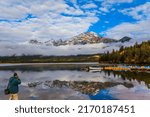  The morning fog rises to the sky. Elderly tourist with a photo bag is photographing Rocky Mountains. Pyramid Mountain is reflected in the Pyramid Lake. The concept of photo tourism