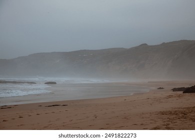 Morning fog shrouds Monte Clérigo beach in Aljezur, Portugal, softening the landscape. Gentle waves lap the shore, creating a serene and mystical coastal scene. - Powered by Shutterstock