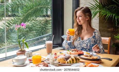 Morning Food. Young Woman Drinks Orange Juice From Glass In Luxury Hotel Restaurant On Dinner Or Lunch. Attractive Female Eat Breakfast, Sitting Next To Window With Tropical Palm Trees. Buffet Food