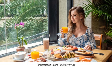 Morning Food. Young Woman Drinks Orange Juice From Glass In Luxury Hotel Restaurant On Dinner Or Lunch. Attractive Female Eat Breakfast, Sitting Next To Window With Tropical Palm Trees. Buffet Food