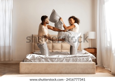 Morning Fun. Playful black husband and wife having fun fighting with pillows and laughing, in domestic bedroom indoor, enjoying carefree pillow battle and flirt together at home interior