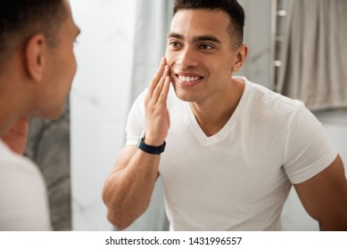 Morning Hygiene Concept. Back Side Close Up Portrait Of Young Happy Satisfied Muscular Guy In White T-shirt Looking At Reflection On Mirror And Touching His Shaving Face