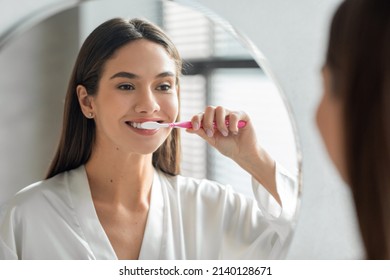Higiene matutina. Retrato De Una Atractiva Joven Que Se Cepa Con Los Dientes Cerca Del Espejo, Una Hermosa Mujer Feliz Que Sonríe A Su Reflexión Mientras Está En El Cuarto De Baño Con cepillo De dientes A Mano, Enfoque Selectivo