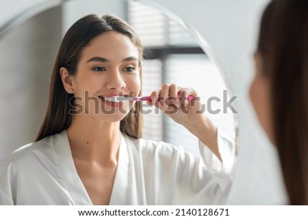 Morning Hygiene. Portrait Of Attractive Young Lady Brushing Teeth Near Mirror, Happy Beautiful Woman Smiling To Her Reflection While Standing In Bathroom With Toothbrush In Hand, Selective Focus Foto stock © 