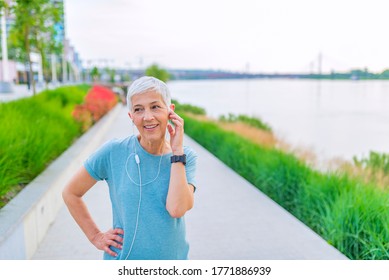 Morning Jogging. A Senior Woman In Her 60s Exercising In The City. She Is Running, Jogging Or Power Walking, Listening To Music Through Earbuds. 