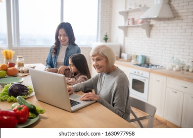 Morning In The Kitchen. Smiling Grandmother Sitting At The Kitchen Table And Looking With Interest At The Laptop, And Next To Mom With A Small Daughter, Making Breakfast, Busy And Joyful.