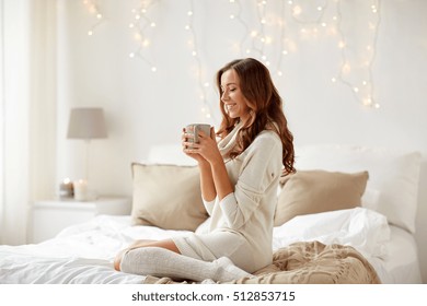 Morning, Leisure, Christmas, Winter And People Concept - Happy Young Woman With Cup Of Coffee Or Tea In Bed At Home Bedroom