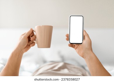 Morning Leisure. Cropped Man Using Smartphone With White Screen While Lying In Bed At Home, Hands Holding Coffee Mug And Blank Cellphone, Shopping Online Or Browsing New App, Mockup Image, POV