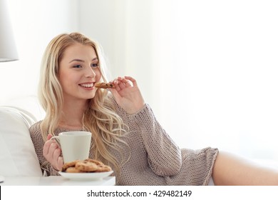 morning, leisure and people concept - happy young woman with cup of coffee or tea eating cookie in bed at home bedroom