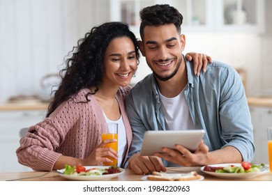 Morning Leisure. Young Happy Arab Spouses Eating Breakfast And Using Digital Tablet In Kitchen, Cheerful Middle Eastern Lovers Spending Time With Tab Computer And Enjoying Tasty Food, Closeup