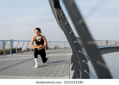 In the morning light, a dedicated young woman performs a lunging stretch on a sleek urban bridge, embracing the freshness of a new day with determination and grace. - Powered by Shutterstock