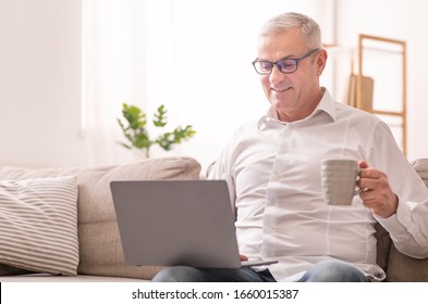 In The Morning. Mature Man Holding Computer On His Lap, Having Tea Sitting On Sofa At Home, Free Space