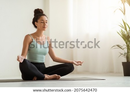 Morning Meditation. Beautiful Calm Woman Meditating At Home In Lotus Position, Peaceful Young Brunette Lady Practicing Yoga, Sitting With Closed Eyes On Fitness Mat In Light Room, Copy Space