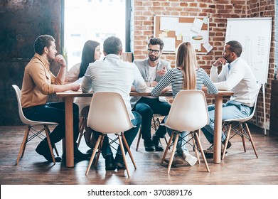 Morning Meeting. Group Of Six Young People Discussing Something While Sitting At The Table In Office Together