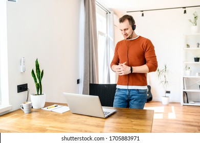 Morning Meeting Via Video Connection. A Guy With Hands Free Headset Is Talking Online Using Laptop, He Stands And Gesturing. Home Office, Online Meeting, Online Conference