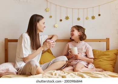 Morning with mom. Young happy mother and cute little daughter wearing pajamas having good talk while drinking tea in bed at home, cheerful older and younger sister spending time together in bedroom - Powered by Shutterstock