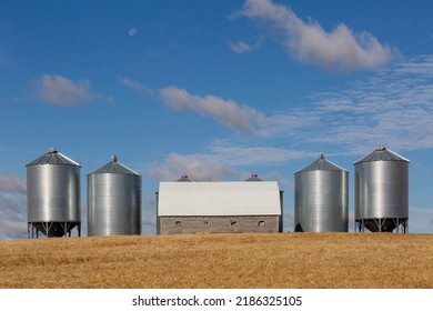 The Morning Moon In A Blue Sky Sets Over Modern, Metal Grain Storage Bins. An Old Wooden Storage Shed Sits Between Them, Slowly Decaying.