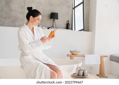 Morning on weekend. Spa treatment and beauty care. Young caucasian woman in bathrobe applying body oil on her skin for softening and moisturizing effect at home.