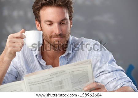 Morning portrait of handsome guy reading newspaper and holding coffee cup.