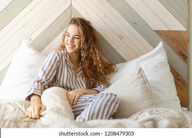 Morning portrait of smiling pretty young woman laughing and looking away. Cheerful girl laying on pillows in striped pyjamas. Happiness and start of new day concept