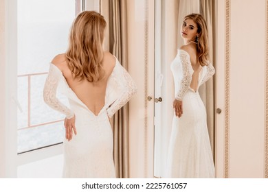 Morning preparation of the bride. Close-up of a bridesmaid, fastening a lot of buttons on the bride's wedding dress. The bride in a white wedding dress with lace standing in the room. Selective focus