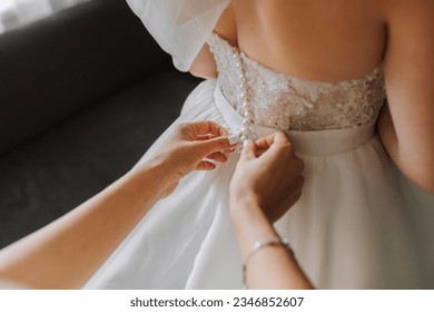 Morning preparation of the bride. Close-up of a bridesmaid's hands fastening the many buttons on the bride's wedding dress. The bride in a white wedding dress is standing in the room.