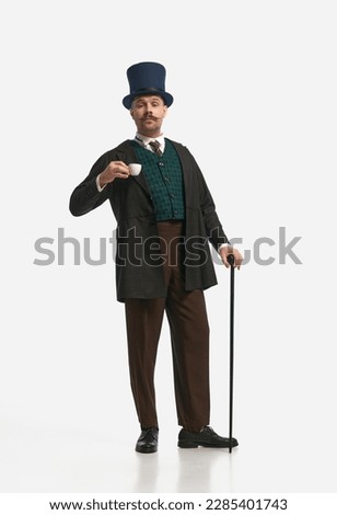 Morning ritual. Portrait of man wearing vintage costume and cylinder hat standing with cane and drinking coffee over white background. Concept of historical remake, comparison of eras, retro, vintage
