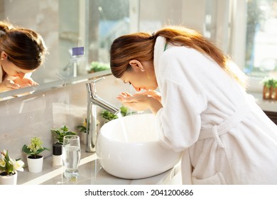 Morning routine. A beautiful woman in a bathrobe washes her face in the bathroom by the sink. Modern interior. Pregnancy. Reflection in the mirror. Clean skin - Shutterstock ID 2041200686