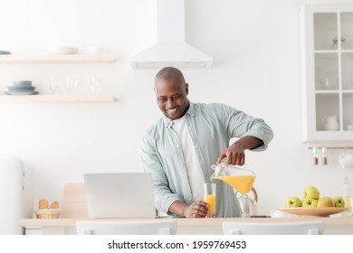 Morning Routine. Happy Mature Black Man Having Video Chat With Family On Laptop Computer, Pouring Juice On The Kitchen Table And Looking At Pc Screen, Empty Space