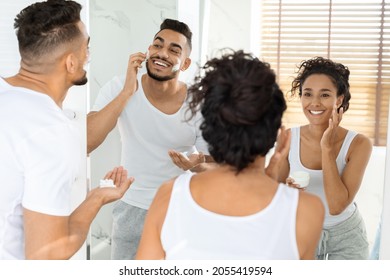 Morning Routine. Happy Young Arab Couple Getting Ready Together Near Mirror In Bathroom, Woman Applying Moisturising Cream While Man Using Shave Foam, Selective Focus On Reflection, Free Space - Powered by Shutterstock