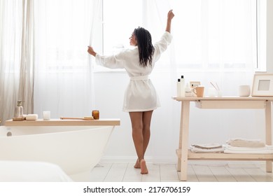 Morning Routine. Rear View Of Happy Black Woman In Silk Robe Opening Curtains In Bathroom, Smiling Young African American Standing Near Big Window, Ready For Beauty Treatments, Free Space