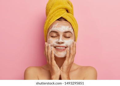 Morning Routine And Relaxing Time Concept. Pleased Young Woman Washes Face, Cleans Skin With Soap, Wears Yellow Towel On Head, Keeps Eyes Shut From Pleasure, Poses With Naked Body. Cleansing Concept