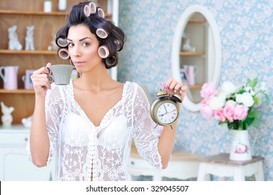 Morning Rush. Beautiful Woman In Hair Curlers Holding Cup Of Coffee In One Hand And A Clock In The Other