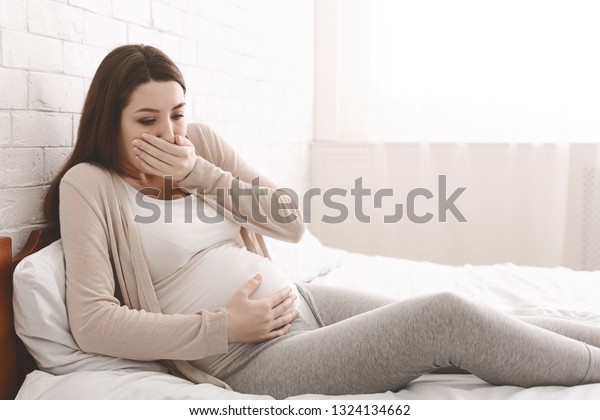 Morning\
sickness. Young pregnant woman sitting on bed, covering her mouth\
feeling nauseous during pregnancy, free\
space