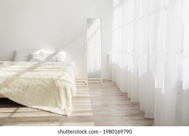 Morning in stylish bedroom in simple minimalist interior. Double bed with pillows, soft blanket, mirror on wooden floor, on white wall background, large window with curtains, nobody, free space