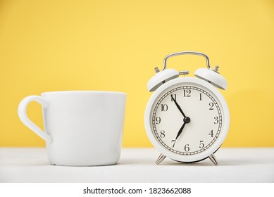Morning time concept. White retro alarm clock and cup on yellow background