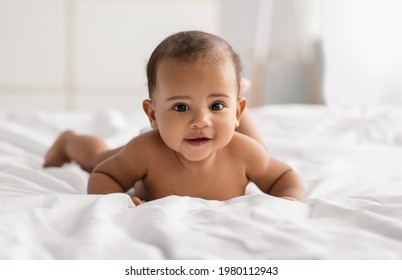 Morning Time. Portrait of cute naked African American baby crawling on bed on the white blanket in bedroom. Smiling black kid lying on tummy, playing, wearing diaper. Free copy space