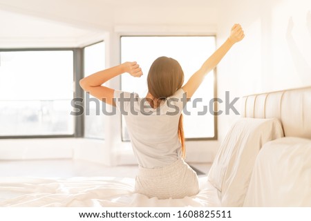 Morning woman waking up in bed in sunrise sunlight glow from window. Early bird happy girl in pajamas on weekend stretching arms. Healthy wake-up routine home lifestyle.