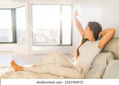 Morning woman waking up stretching in bed in sunshine sunrise early bird happy Asian girl in pajamas on weekend in sun glow. Healthy wake-up routine home lifestyle.
