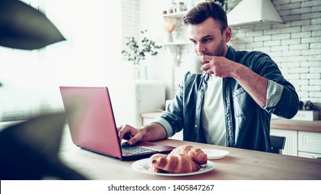 Morning Work With Beverage. Happy Smiling Man Is Using Laptop And Drinks Coffee Or Tea While Sitting On The Kitchen At Home