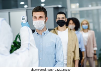 Morning at work during covid-19 epidemic. Person with contactless thermometer measures temperature of multiracial employees in protective masks an entrance of modern office, copy space - Powered by Shutterstock