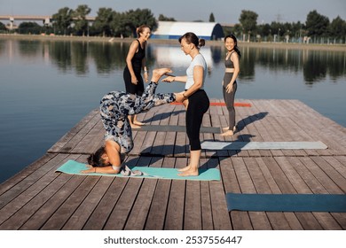 Morning yoga class on a wooden pier in the city. Young women stand in a headstand on mats. The instructor helps to do the exercise. A group of women are doing stretching or pilates outdoors. - Powered by Shutterstock