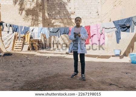 Moroccan girl dressed in a white school gown plays with a spinning top in a small square with dirt floor and clothes hanging