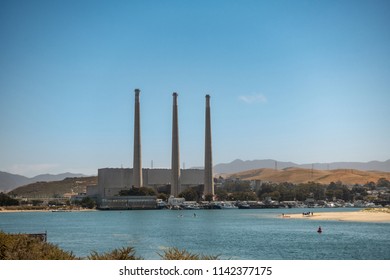 Morro Bay, California, USA - July 10, 2018: Closed Dynergy gas power plant in port under blue sky with brown hills in the back. Three smoke stacks are called Three Fingers. Estuary. outlet to ocean.