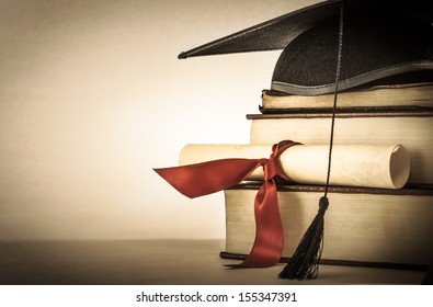A mortarboard and graduation scroll, tied with red ribbon, on a stack of old battered book with empty space to the left.  Slightly undersaturated with vignette for vintage effect.