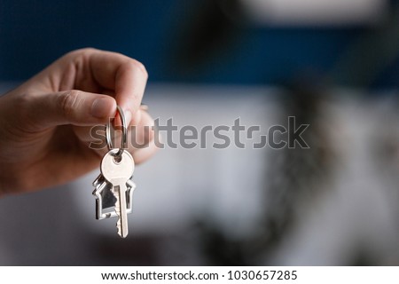 Mortgage concept. Men hand holding key with house shaped keychain. Modern light lobby interior. Real estate, moving home or renting property.