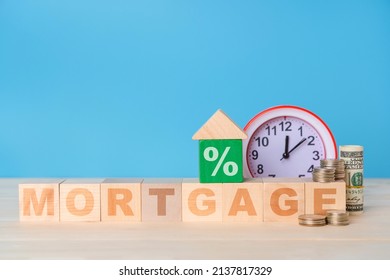 Mortgage Concept. The Word Mortgage On Wooden Cubes, Next To Dollar Bills, Coins And An Alarm Clock. Toy Wooden House With A Percent Sign On A Blue Background, Copy Space