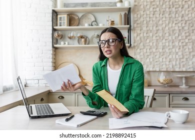 Mortgage Loan. Upset And Angry Young Woman Holding A Rent Bill In Her Hands. Received A Letter About The Debt. Sittin G In The Kitchen At Home At The Table With A Laptop In Glasses And A Green Jacket